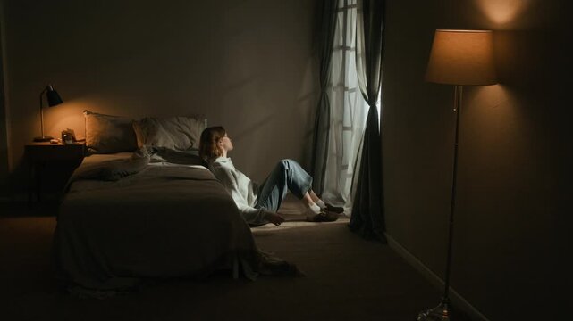 Long stab shot of gen Z Caucasian girl lost in thoughts sitting on floor by bed and window after sleepless night in low key room