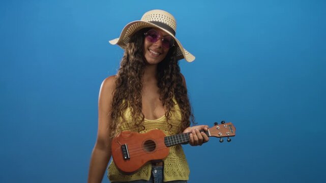 Woman strumming ukulele in studio wearing wide sun hat and purple sunglasses, holding and motioning with raised right hand while smiling; playful summer fun.