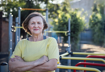 Senior woman with short hair wearing yellow shirt stands confidently with arms crossed in outdoor fitness area with exercise equipment in the background © Dasha Petrenko