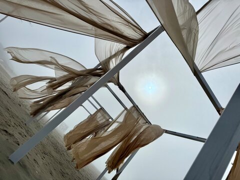 Tilted view of metal beach frames with flowing translucent fabric in foggy coastal light, creating an artistic seaside installation. Liepaja