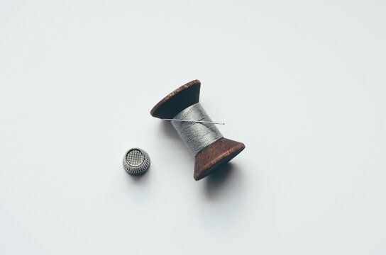 Minimalist top view of vintage wooden spool with silver thread, needle, and thimble on white, ideal for tailoring and craft advertising.
