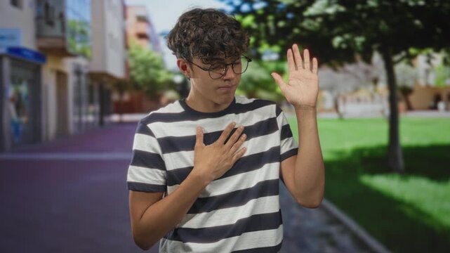 Teenage boy wearing glasses and a striped shirt places hand on chest and raises other hand on street; sincerity pledge.