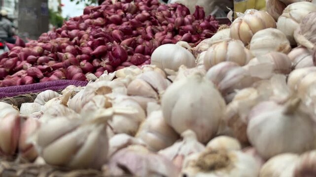A wide selection of garlic heads lies across a vendor table, with heaps of red onions and active market trade forming the background. Wholesale vegetables.