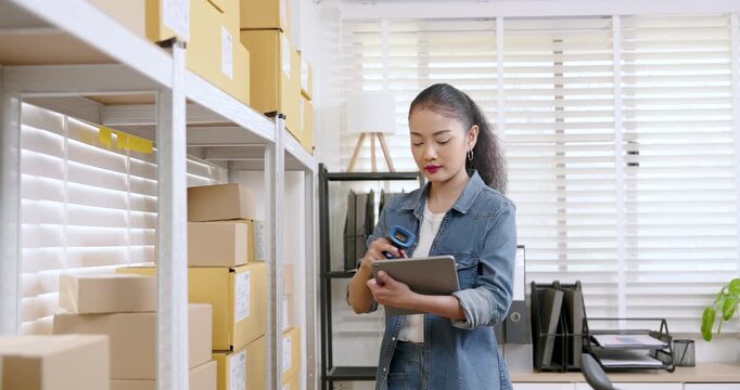 Asian woman small business owner slowly reviews inventory list on tablet while standing between warehouse shelves filled with online shopping parcel boxes ready for shipment
