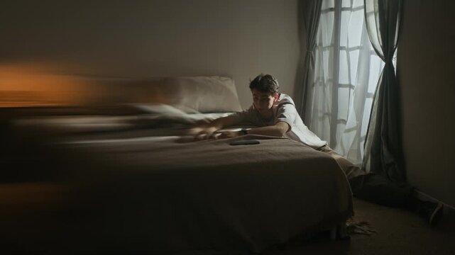 Long motion blur of apathetic young Caucasian man sitting on floor by bed and window, checking smartphone for notifications in lowkey bedroom