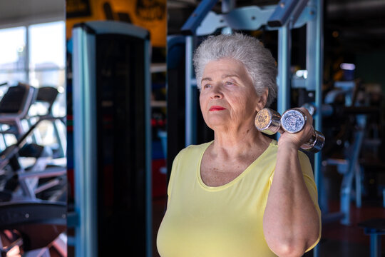 Senior woman in her mid-seventies with silver gray hair wearing yellow athletic tank top performing dumbbell bicep curl exercise with concentrated upward gaze at modern fitness center.  