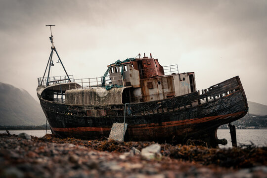 View of the decaying shipwreck, a relic of time, rests on the shore against a backdrop of moody skies and distant mountains, Fort William, Scotland, United Kingdom.