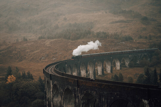View of a vintage steam train puffing white clouds as it traverses the Glenfinnan Viaduct, a testament to engineering amidst the misty highlands, Glenfinnan, Scotland, United Kingdom.