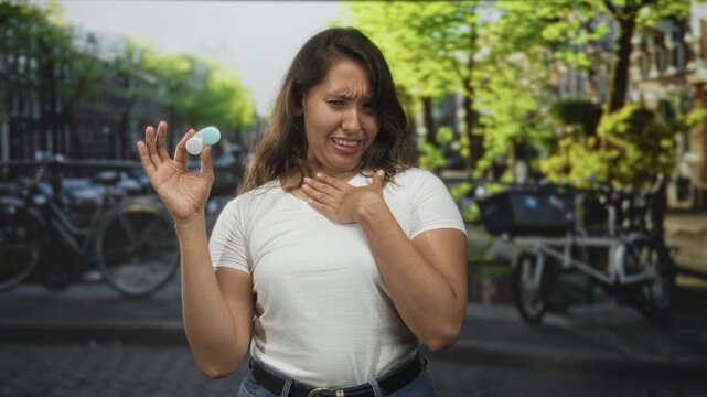 Woman holding a contact lens case with one hand and touching chest with the other on a street in amsterdam; disgust hygiene concern.