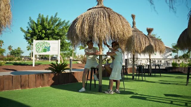 Affectionate family enjoy sunny day outdoors under straw tiki umbrella, with mother hugging and caressing young daughter while father watches lovingly. Family, love, parenting, and lifestyle content.