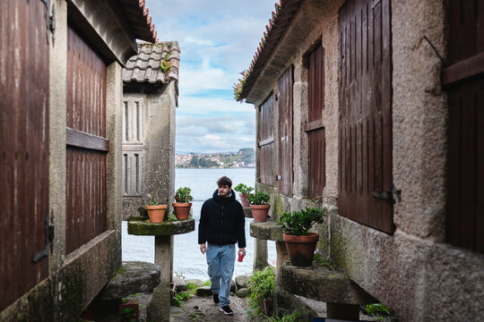 Young man exploring Combarro's historic fishing village, walking a narrow stone path between ancient horreos toward the scenic coastal ria, summer travel atmosphere
