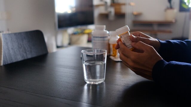 Person holding a prescription medicine bottle and reading label for dosage or instructions, preparing to take medication with a glass of water on a table at home