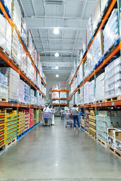 A wide interior photograph of a large warehouse-style wholesale shopping center featuring long aisles lined with tall pallet racks filled with bulk merchandise. The image captures the spacious retail 