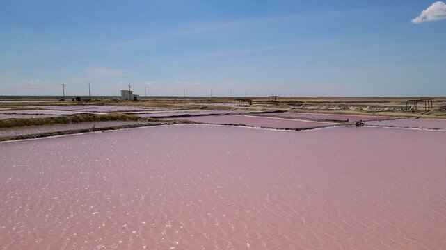 Vibrant pink salt ponds spread across the land, contrasting with the blue sky. Equipment is visible in the distance.