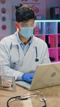 Likable confident experienced male chemist in white coat and medical mask,gloves and eyewear entering results of chemical experiemtns with liquids into computer in dark lighted workroom