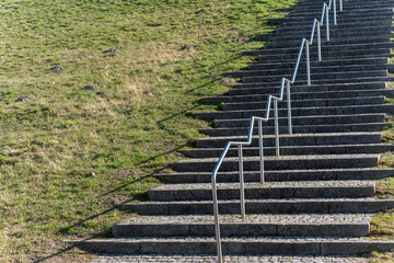 Sunlit stone steps climb toward grassy hillside. Metal railings line each stair for safety. Shadows cast by sun create contrast. Grass patches show natural wear and tear