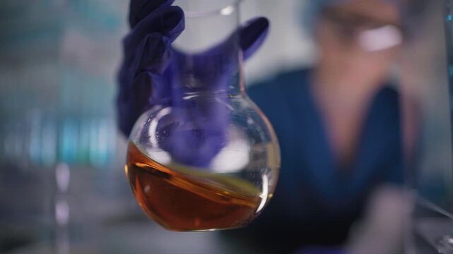 Female researcher preparing a new type of enriched bio fuel. Holding a glass flask with orange liquid close to the camera. Selective focus