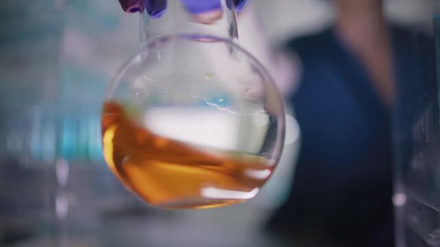 Female researcher preparing a new type of enriched fuel. Holding a glass flask with orange liquid close to the camera and swirling the liquid. Selective focus