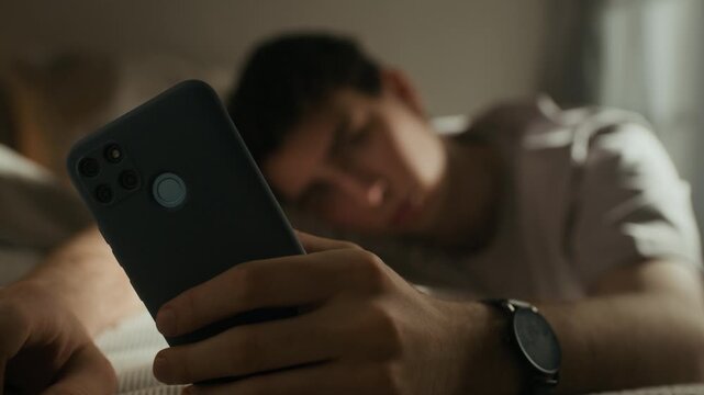 Rack focus of gen Z Caucasian man resting in bed with smartphone and doomscrolling while feeling unhappy in dimly lit bedroom