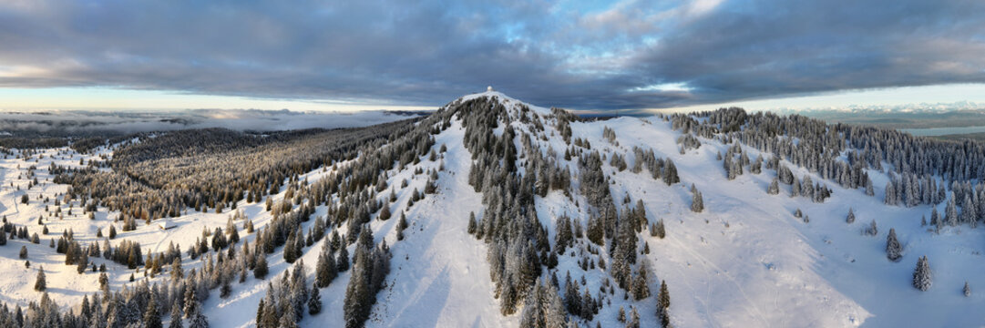 Panoramic aerial view of La Dole snow-covered mountain peak and lush forests under a dramatic sky, crowned by a solitary structure at the summit, Gingins, Vaud, Switzerland.