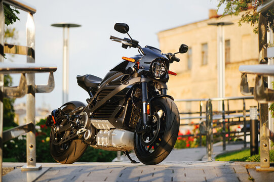 A horizontal wide shot of a modern black electric motorcycle with orange turn signals, parked on a stone walkway with a blurred historic building in the background.