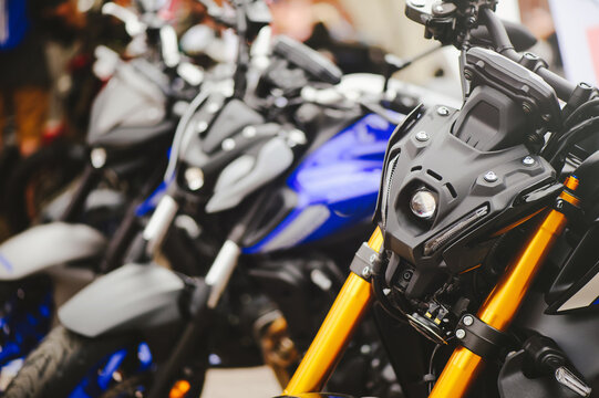 A horizontal shot focusing on the futuristic LED headlight assembly and golden front forks of a modern street motorcycle, with other bikes blurred in the background.
