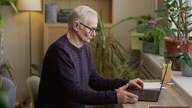 Side view slowmo shot of senior Caucasian man in eyeglasses and casualwear sitting at desk in front of laptop and gesticulating expressively while talking to web camera, working in cozy green office