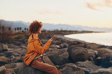 Woman with curly hair wearing orange jacket sitting on rocks by the sea, smiling while holding...