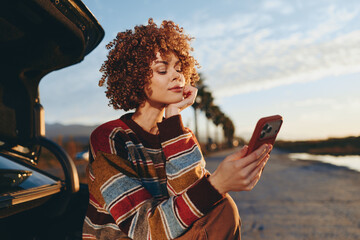 Woman with curly hair wearing rainbow sweater sitting outdoors near car, smiling while looking at...