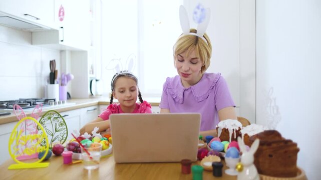 Mother and daughter wearing bunny ears celebrate Easter virtually with a video call, surrounded by decorated eggs and traditional cakes