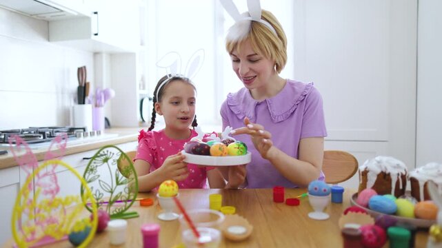 A mother and her daughter wearing bunny ears are happily decorating Easter eggs in a bright kitchen, surrounded by festive decorations and cakes