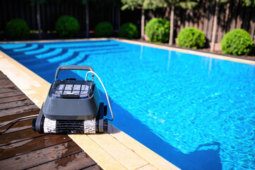 Naklejka premium A pool cleaning robot is placed on the edge of an outdoor swimming pool with clear blue water. Green bushes and trees in the background create a cozy private backyard atmosphere