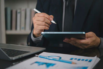 Fototapeta premium businessman in suit using digital tablet and stylus pen with financial graph and laptop on dark desk. Corporate business, executive manager, data analysis and digital technology concept.