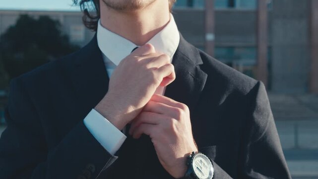 Young man in a stylish business suit and a white shirt walks down the street and adjusts his tie, slow motion. A businessman tightens his tie while walking outdoors, soft focus. Business concept