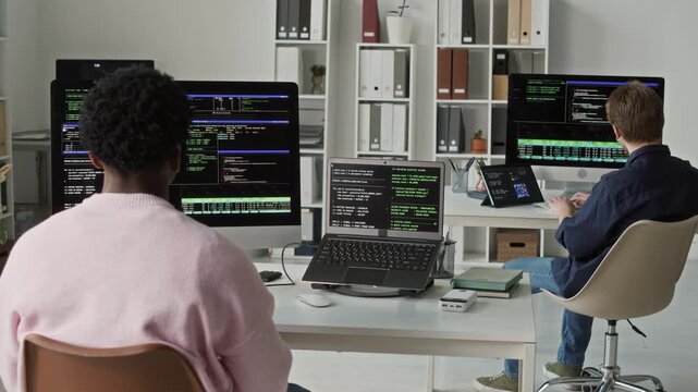 Side view of focused young adult Black male application developer multitasking between laptop and desktop computer at coding desk while typing on keyboards in modern office