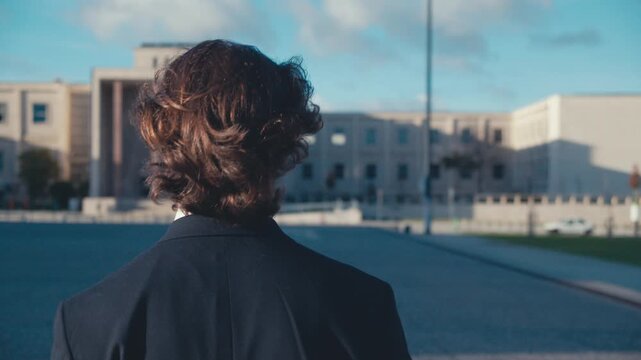 Rear view, close-up of a young man with flowing hair in the wind in a stylish classic business suit, who is walking alone through a deserted city. Slow motion.