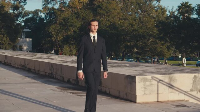 Stylish young man with medium-length hair in an elegant classic business suit and tie walks in a city during sunset. Slow motion.
