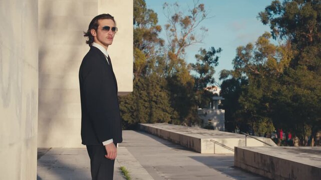 A young confident man in golden sunglasses in a stylish classic business suit with a tie comes out from behind the wall and looks into the distance. Slow motion.