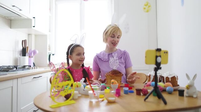 A mother and daughter wearing bunny ears are live streaming their Easter baking activities on a smartphone, surrounded by decorated eggs and cakes