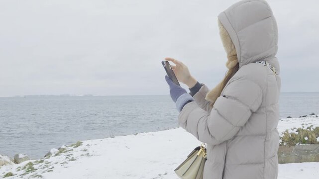 A woman dressed in a warm winter coat stands by the snow-covered shoreline, using her mobile phone. The serene environment and winter scenery create a peaceful atmosphere, perfect for a leisurely stro