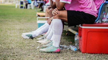 Soccer players resting on the team bench during a football match, wearing pink jerseys and white socks, with water bottles and cooler box nearby, showing break time, teamwork, and sports recovery © pixfly