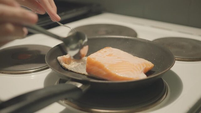 A close-up view of salmon being cooked in a frying pan, showcasing the sizzling moment. A hand is seen using a spatula to flip the fish, highlighting the cooking process in a kitchen environment.