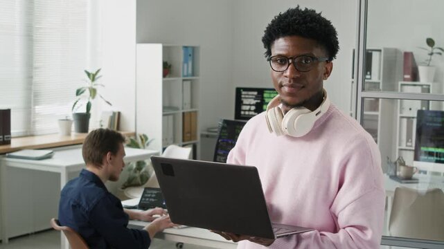 Medium portrait of young adult African American developer wearing headphones over neck and holding laptop smiling at camera while standing in modern IT office with open plan interior, slow motion