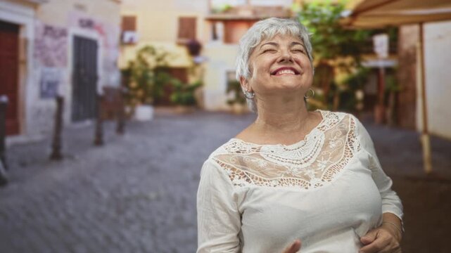 Woman with hands raised smiling and fist pumping, laughing in a white lace blouse on a cobblestone street in a historic town, grey hair visible; joy freedom.