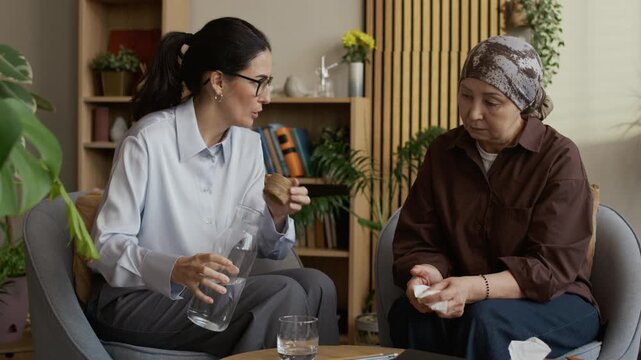 Medium long shot of middle-aged Caucasian female counselor offering glass of water to mature Asian female patient wearing headscarf and gently comforting her by shoulder during office consultation