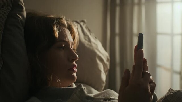 Close up lowkey shot of disinterested gen Z woman doomscrolling on mobile phone while resting in bed experiencing insomnia at night