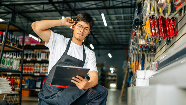 Young man wearing apron and glasses checking inventory on a clipboard in a hardware store