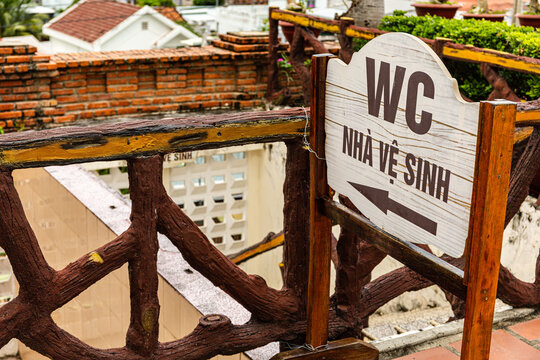 WC restroom sign with Vietnamese 'Nh&agrave; Vệ Sinh' (lavatory)  text mounted on a rustic wooden railing, pointing the way to a public toilet in a tropical outdoor travel setting,