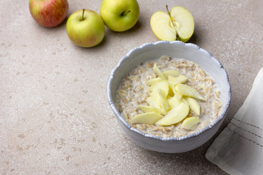 Oatmeal porridge with milk topped with fresh apple slices in ceramic bowl. Nutritious breakfast with oats and fruit on light kitchen table