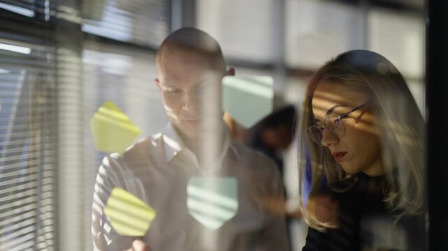 Business professionals collaborate on ideas using colorful sticky notes affixed to a glass surface. Colleagues are brainstorming and planning solutions together in a modern office.